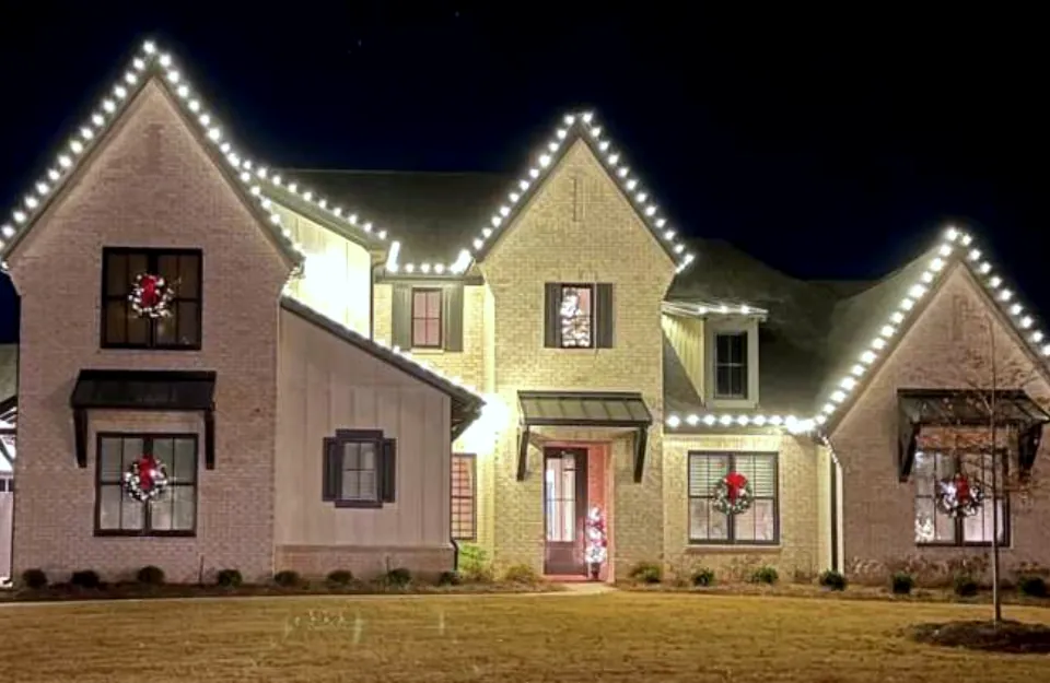 Light brick house with a tall front gable lit end to end and wreaths in the upstairs windows.