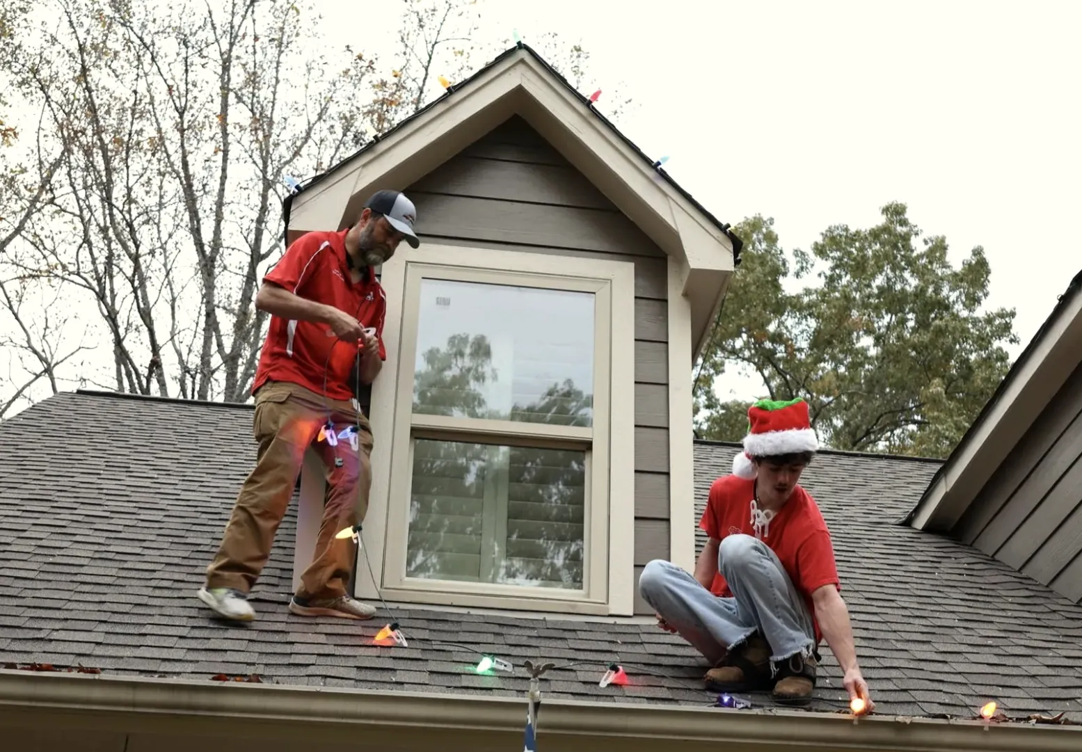 Two crew members on a shingle roof beside a dormer, working a light strand along the edge.