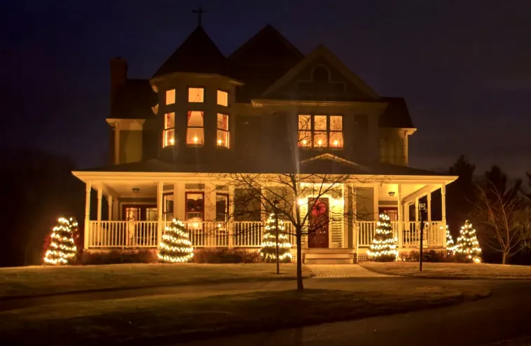 Victorian-style house with a wraparound porch, glowing windows and small lit trees in the yard.